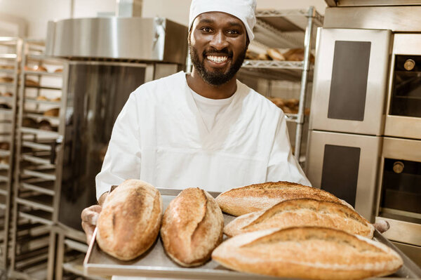 handsome african american baker with tray of fresh loaves of bread on baking manufacture