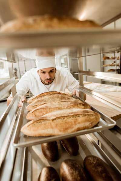 handsome baker putting trays of fresh bread on stand at baking manufacture