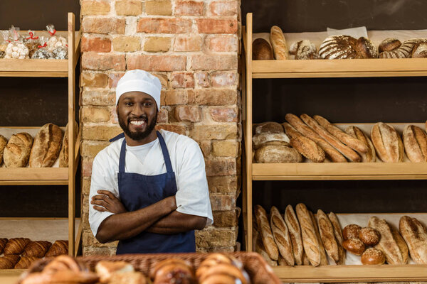 handsome smiling baker with folded arms standing at pastry store