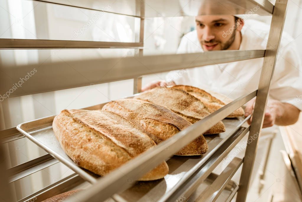 close-up shot of handsome baker putting trays of fresh bread on stand at baking manufacture