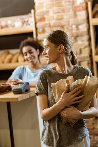 happy young woman leaving pastry store with various bread in paper bags