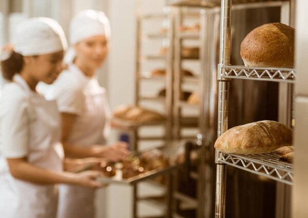 bakers working at baking manufacture with fresh loaves of bread lying on shelves on foreground