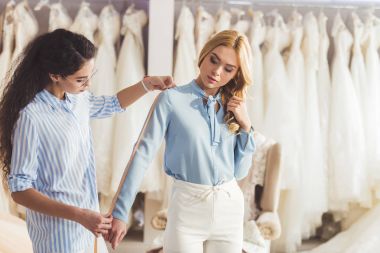 Young woman and professional tailor taking measurements in wedding fashion shop clipart