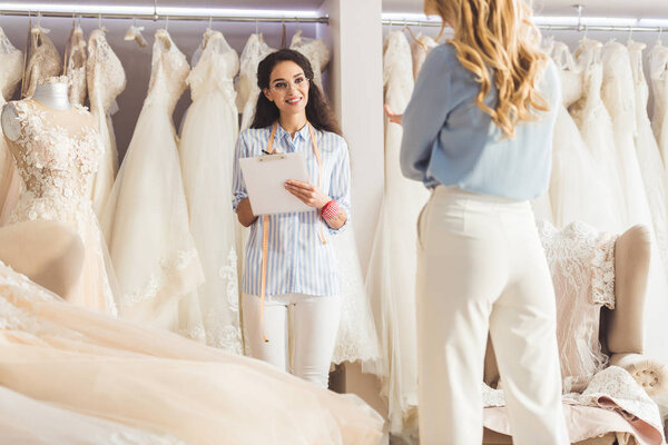 Beautiful bride and female tailor discussing dress design in wedding salon