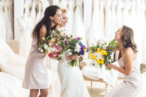 Young bride and bridesmaids holding tender flowers bouquets in wedding atelier