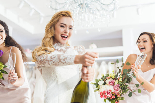Young smiling bride and bridesmaids opening champagne in wedding fashion shop