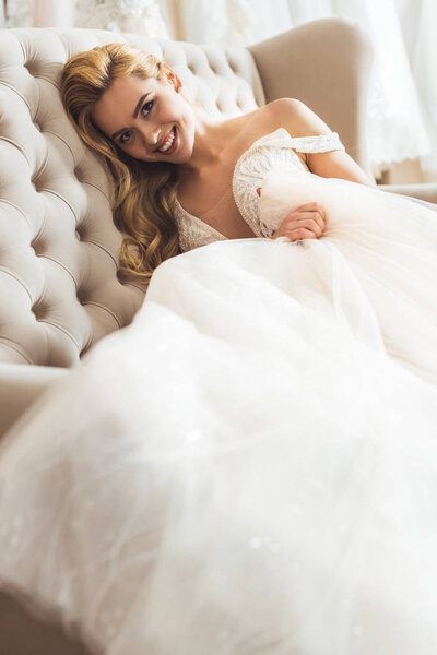 Smiling bride wearing tulle dress sitting on sofa in wedding atelier