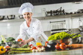 lächelnd attraktiver Koch nimmt Schüssel mit Gemüse und blickt in die Kamera der Restaurantküche