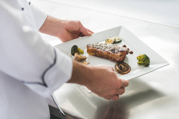 cropped image of chef holding plate with cooked steak and vegetables at restaurant kitchen