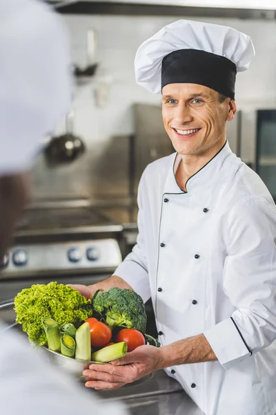 Sonriente Chef Sosteniendo Tazón Con Verduras Mirando Colega Cocina Del —  Fotos de Stock