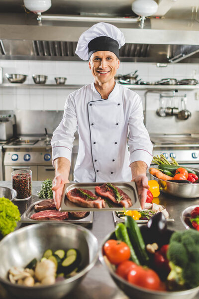 smiling chef taking tray with uncooked meat at restaurant kitchen