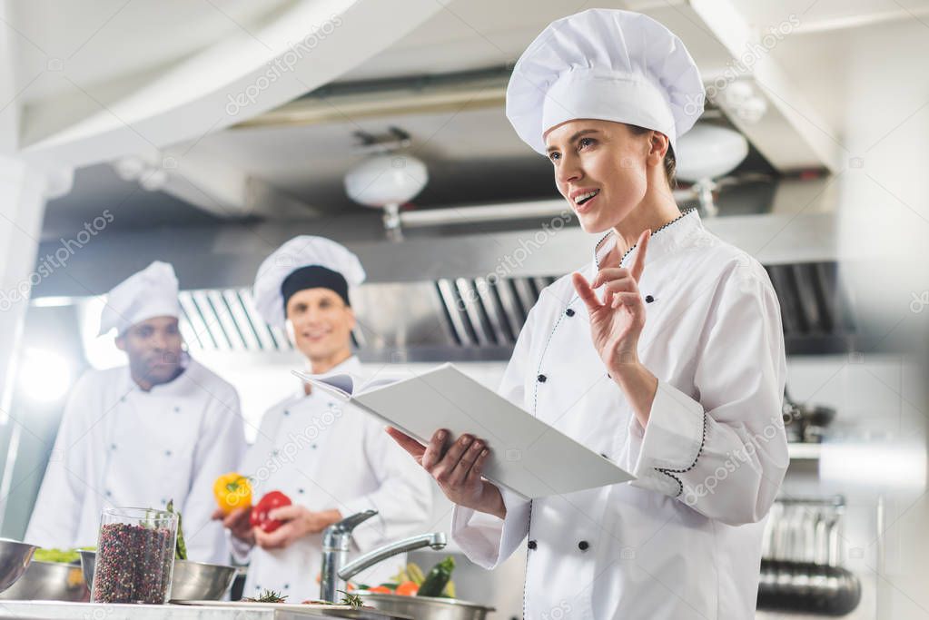 attractive chef holding recipe book and showing idea gesture at restaurant kitchen