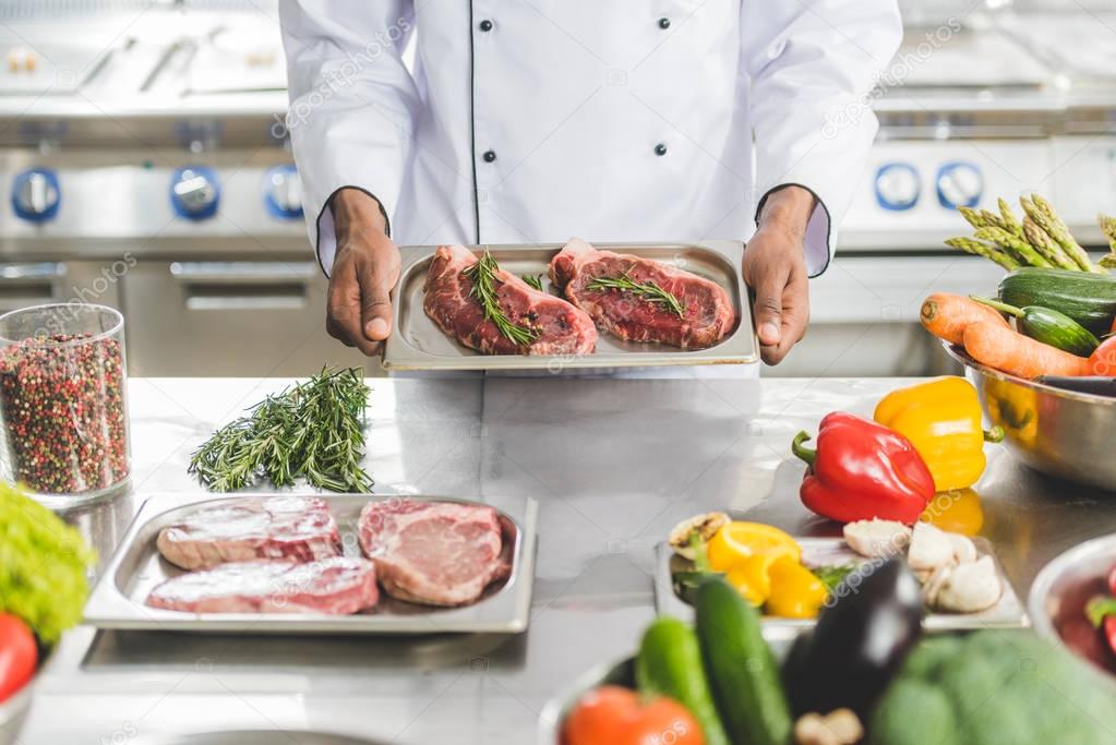 cropped image of african american chef holding tray with uncooked meat at restaurant kitchen