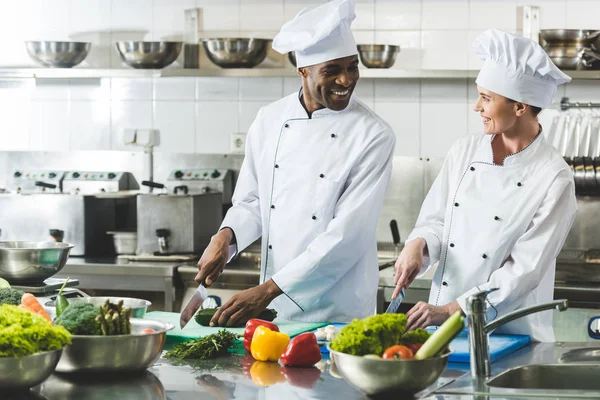 Chefs Multiculturales Sonrientes Cortando Verduras Cocina Del Restaurante Mirándose —  Fotos de Stock