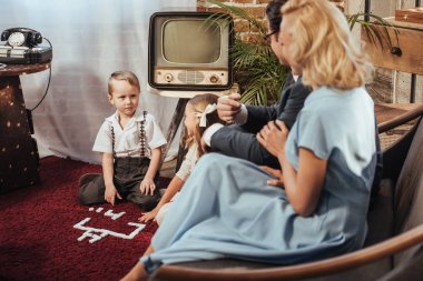 parents sitting on sofa and looking at adorable children playing at home, 50s style   clipart