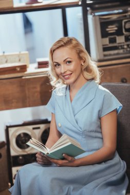 beautiful blonde woman in blue dress holding book and smiling at camera, 50s style clipart