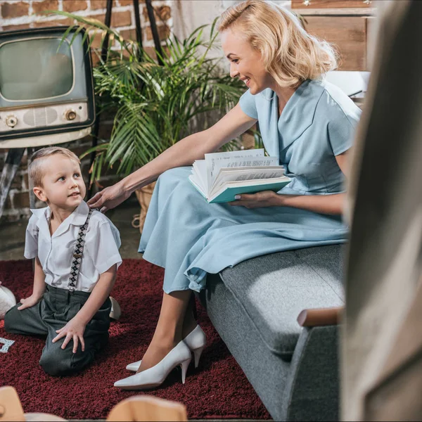 Feliz Mujer Rubia Leyendo Libro Mirando Lindo Pequeño Hijo Jugando — Foto de Stock