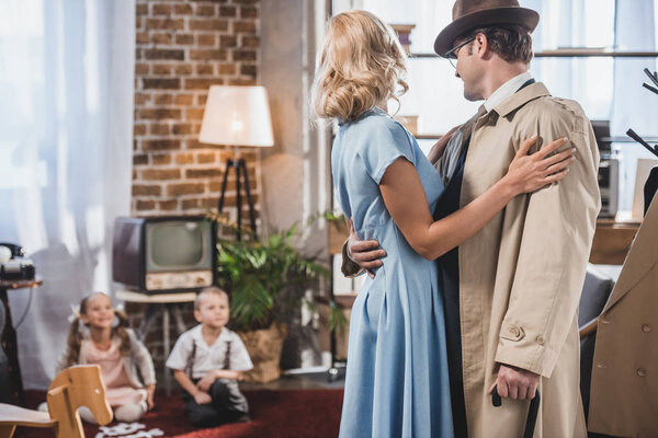 счастливые родители обнимаются и смотрят на милых маленьких детей, играющих дома, стиль пятидесятых
