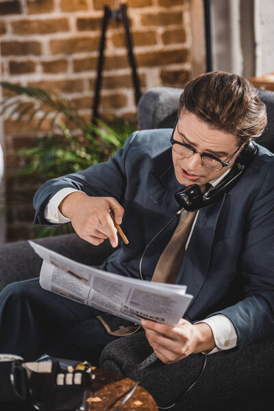 emotional man in suit and eyeglasses pointing at newspaper and talking by vintage telephone