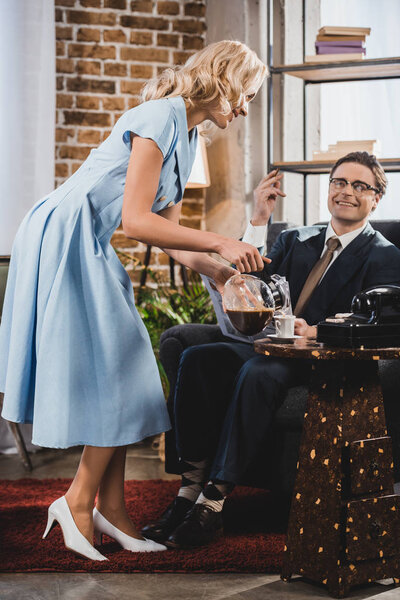 smiling woman pouring coffee to happy husband in suit and eyeglasses sitting in armchair, 1950s style