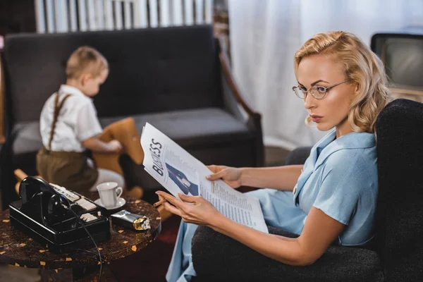 Mujer Gafas Lectura Periódico Negocios Mientras Que Pequeño Hijo Sentado — Foto de Stock