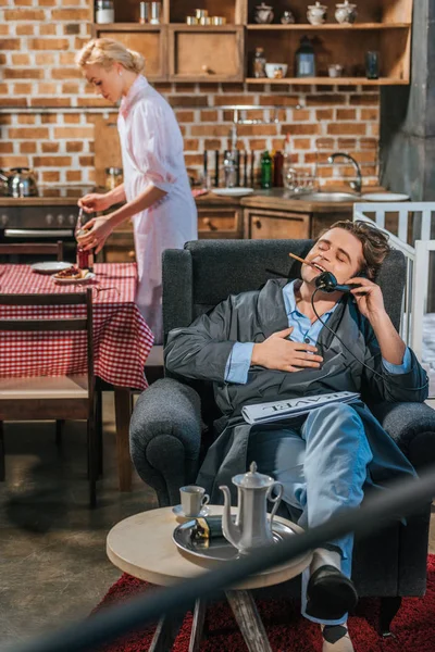 smiling man in robe smoking cigarette and talking by vintage telephone while wife preparing breakfast behind