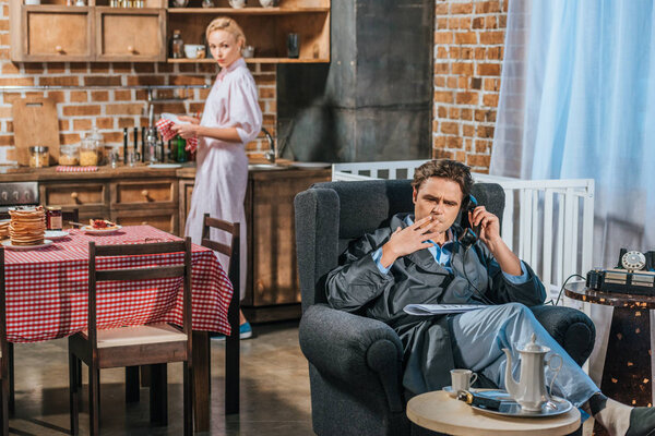 Мужчина в халате разговаривает по старинному телефону и курит сигарету, пока жена стоит позади на кухне
