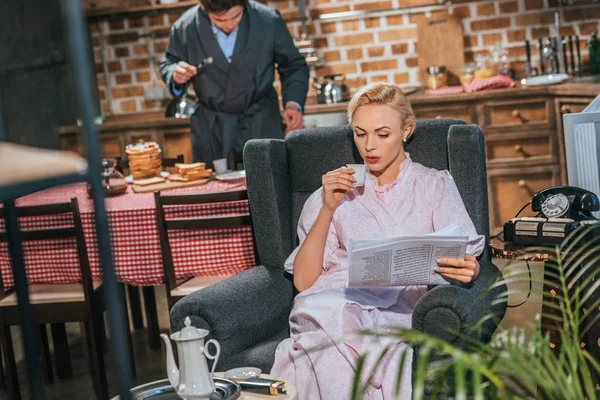 Mujer Vestida Bebiendo Café Leyendo Periódico Mientras Marido Prepara Desayuno — Foto de Stock