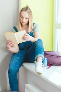 Kitap okuma ve pencere pervazına soda ve sırt çantası ile oturan genç kız öğrenci