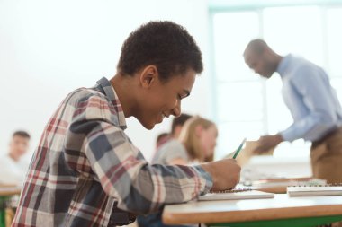 Ders kitabı öğretmen ve sınıf arkadaşları ile yazma gülümseyen Afro-Amerikan çocuk yan görünüm açık arka plan 