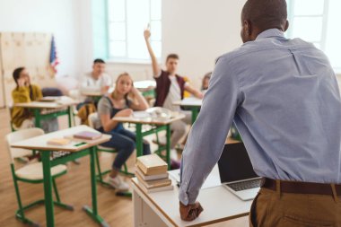 Rear view of african american teacher and schoolboy with arm up in classroom 