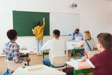 Kız öğrenci öğretmen sınıfta oturma ile tebeşir tahtası ve sınıf yazılı arka görünümü 