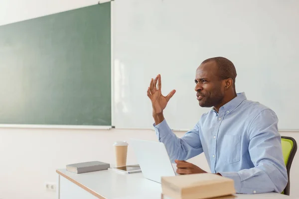 不満をアフリカ系アメリカ人教師ジェスチャーとノート パソコンと机に座って  — 無料ストックフォト