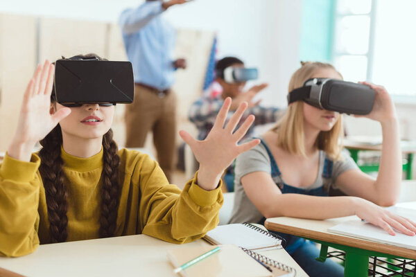Две школьницы-подростки используют гарнитуры виртуальной реальности и одноклассницу с учителем позади
 