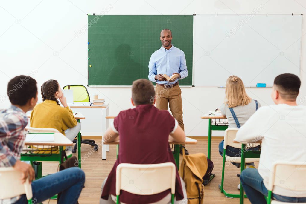 Rear view of high school teenage students listening african american teacher with book in classroom 