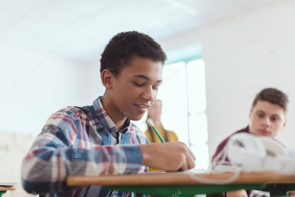 Low angle view of african american teenage high school student writing in textbook and classmates behind 