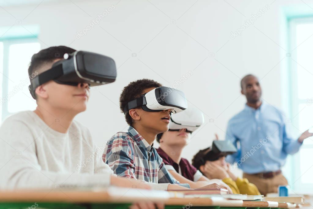 Multicultural schoolchildren using virtual reality headsets and african american teacher standing behind