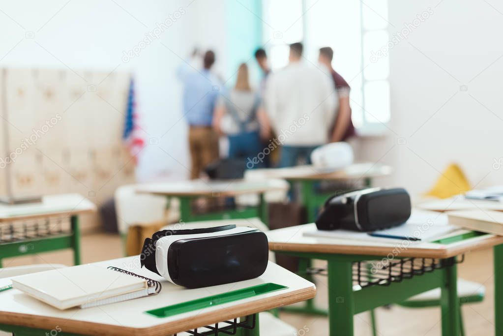 Virtual reality headsets on tables with teacher and high school students standing behind in classroom