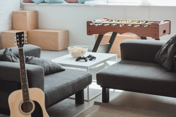 interior of modern living room with guitar, armchairs, table football and table with joysticks and popcorn 