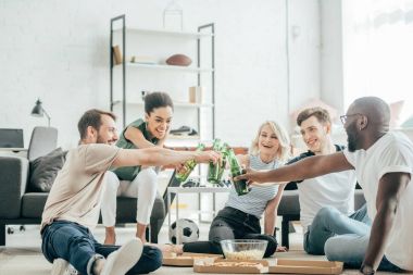 happy multiethnic friends sitting on floor and clinking with bottles of beer  clipart