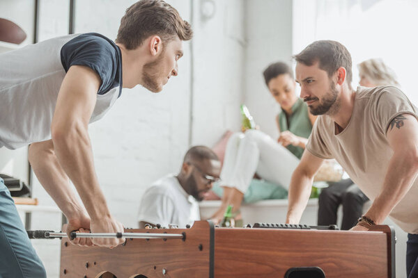 young men with facial expression playing table football with friends sitting behind 