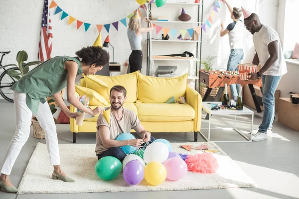 パーティー ハットを風船で床と友人と女性の笑みを浮かべて男に提案している若いアフリカ系アメリカ人女性 — ストック写真