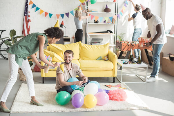 young african american woman proposing party hat to smiling man on floor with balloons and woman with friend 