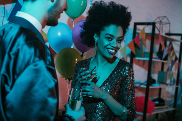 African american woman holding glass with cocktail and talking to man at party