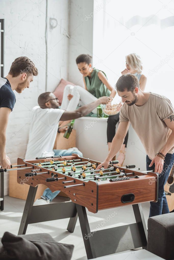  two young men playing table football while their friends watching with beer bottles and popcorn