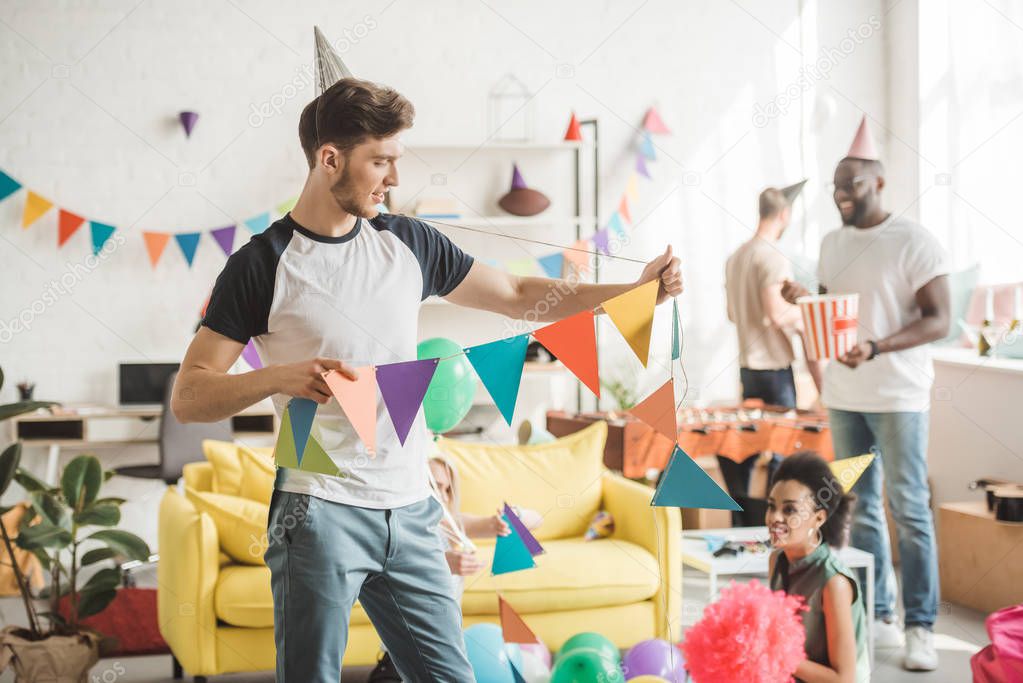 young man in party hat holding string with party garlands and friends standing behind in room