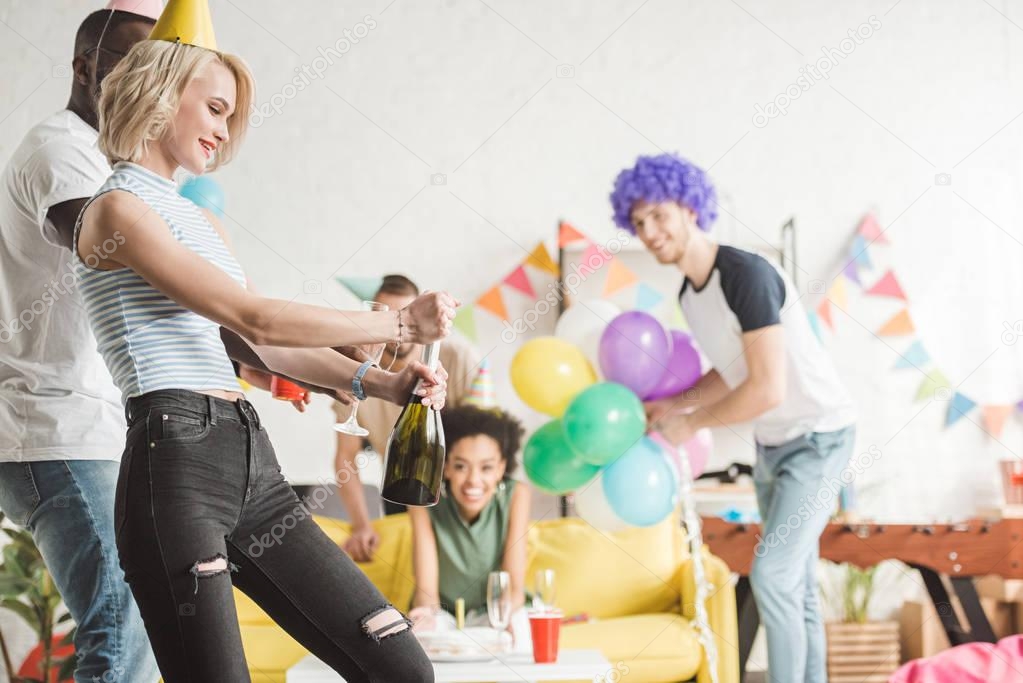 Blonde woman opening champagne in front of partying friends