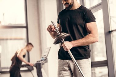 cropped image of sportsman fixing weight plates on iron bar for training in gym clipart