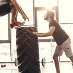 Side view of muscular young men in sportswear training with tire in gym