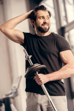 smiling sportsman holding iron bar and touching hair in gym clipart